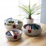 Three colorful woven baskets holding various items, including yarn, a notebook, keys, a wallet, and a smartphone, placed on a wooden table next to a potted plant.