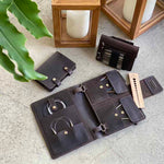 Leather knitting needle organizer set with circular needles, storage pouches, and measuring tool, displayed on a concrete surface near wooden candle holders and a green plant.
