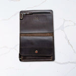 Dark leather tablet case with a button closure and slot for stylus, placed on a white marble surface.