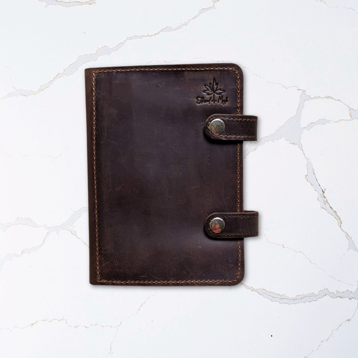 Dark brown leather notebook cover with strap closures placed on a white marble surface.