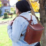 Woman wearing a denim jacket and a black beanie, carrying a brown leather backpack, standing next to a tree in a park during autumn.