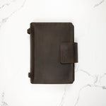 Dark leather notebook cover with snap closure on a white marble background.
