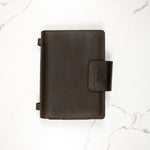 Dark brown leather binder with logo on clasp, placed on a white marble surface.