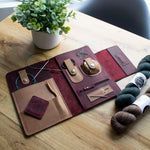 Leather knitting organizer with tools and yarn on a wooden table next to a small potted plant.