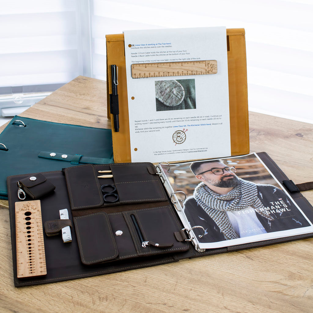 Knitting pattern folders with accessories including scissors, measuring tape, and crochet hooks on a wooden table.
