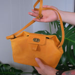 Person holding a small yellow leather handbag with a strap and brass buckle in front of a green leaf background.