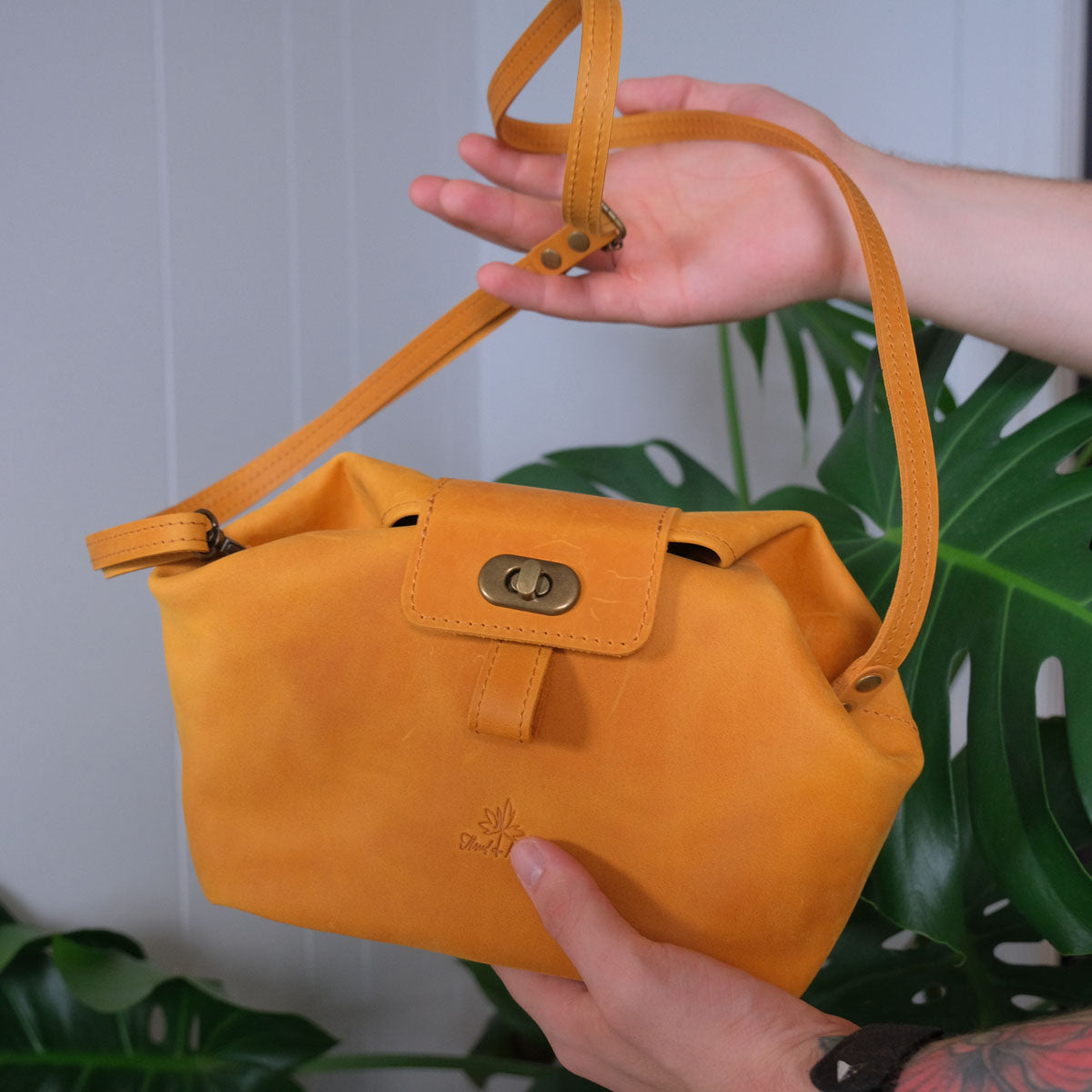 Person holding a small yellow leather handbag with a strap and brass buckle in front of a green leaf background.